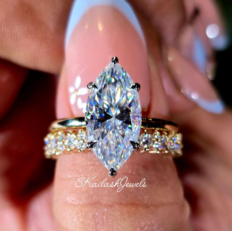 May include: Marquise-cut diamond engagement ring with a gold band and a smaller diamond band. The main diamond is set with four prongs. The ring is on a finger with a light pink manicure. The text "SKailashJewels" is visible.