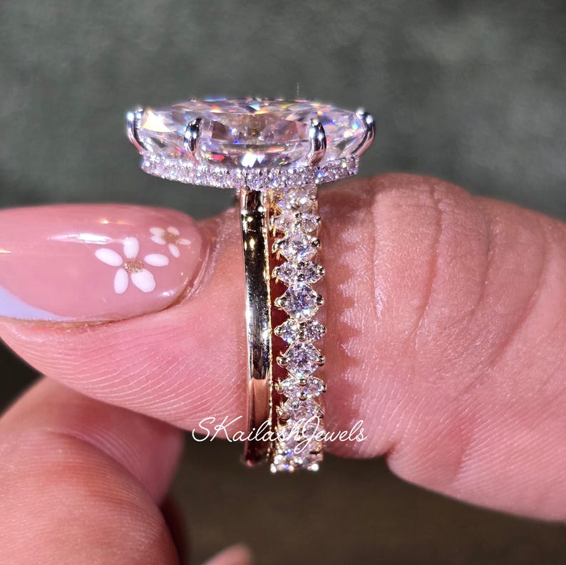 May include: A close-up of a diamond engagement ring with a large oval-cut center stone and a halo of smaller diamonds. The band is gold, and a diamond eternity band sits next to it. The text "Skaila Jewels" is visible.