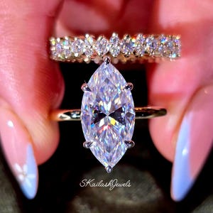 May include: Two gold rings are displayed. The primary ring features a large, clear, marquise-cut gemstone. A second ring, set with multiple smaller, clear gemstones, sits behind the first. The text "SkailashJewels" is visible.