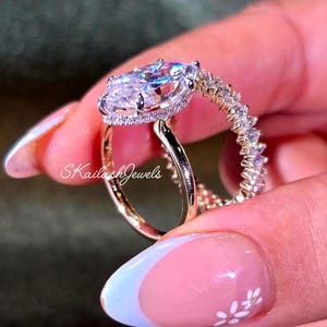 May include: A gold engagement ring featuring a large, clear, pear-shaped gemstone. The band is adorned with smaller, sparkling stones. The ring is held up, showcasing the intricate design and the text "SkailashJewels".