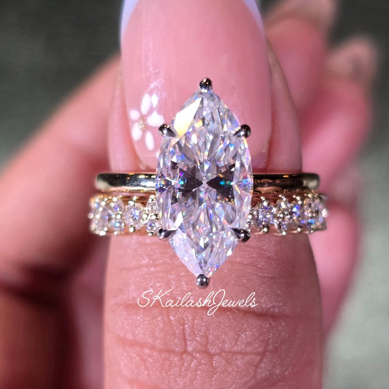 May include: A marquise-cut diamond engagement ring with a gold band, set with smaller diamonds. The ring is paired with a gold band with small diamonds. The text "SKailashJewels" is visible.