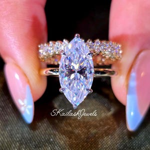 May include: A marquise-cut diamond engagement ring with a gold band, paired with a gold band ring with small diamonds. The rings are held up, with the text "SkailashJewels" visible.