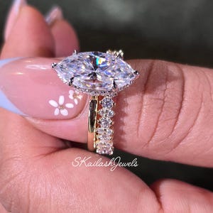 May include: A marquise-cut diamond engagement ring with a halo setting and a diamond band. The ring is held up by a hand with a floral nail design. The text "SKailashJewels" is visible.
