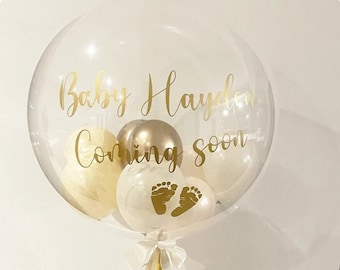 May include: A clear balloon with a gold ribbon and smaller balloons inside. The balloon has the text "Baby Hayden Coming Soon" written on it in gold lettering. The smaller balloons are white and gold.