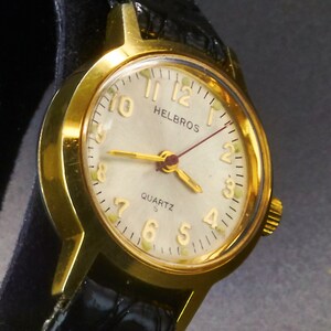May include: A gold-toned Helbros quartz watch with a cream-colored face, gold hands, and black leather band. The watch face features gold numerals and the word "QUARTZ". The watch is a classic timepiece.
