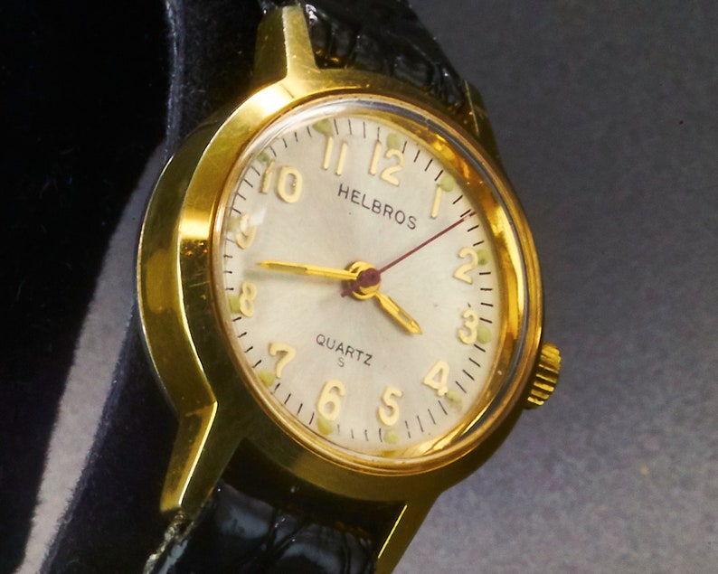 May include: A gold-toned Helbros quartz watch with a cream-colored face, gold hands, and black leather band. The watch face features gold numerals and the word "QUARTZ". The watch is a classic timepiece.
