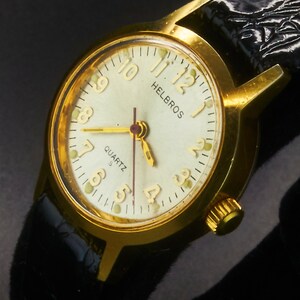 May include: A gold-toned Helbros quartz watch with a cream-colored face and black leather band. The watch face displays the time with gold hands and numerals. The watch has a gold crown and a black leather strap.