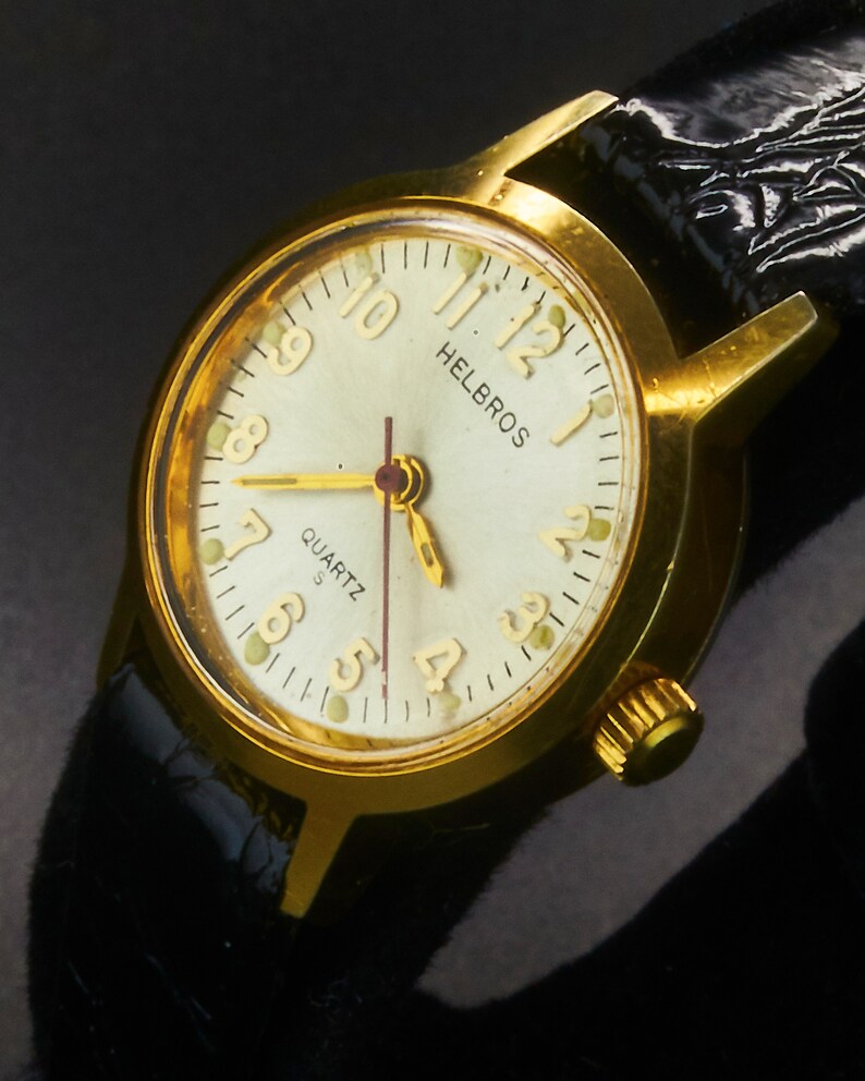 May include: A gold-toned Helbros quartz watch with a cream-colored face and black leather band. The watch face displays the time with gold hands and numerals. The watch has a gold crown and a black leather strap.