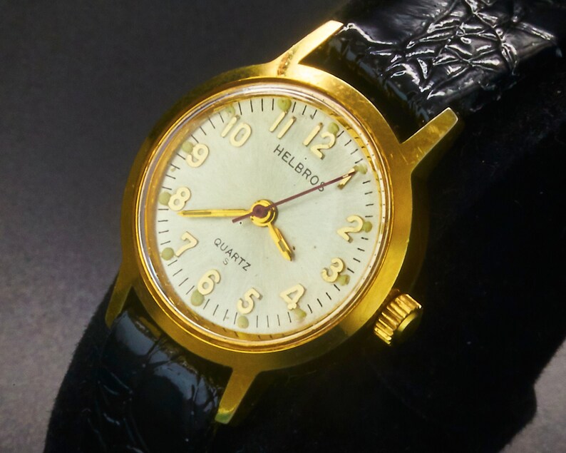 May include: A gold-toned Helbros Quartz watch with a light green face and black leather band. The watch face features gold hands and numerals. The watch is a classic design, suitable for everyday wear.