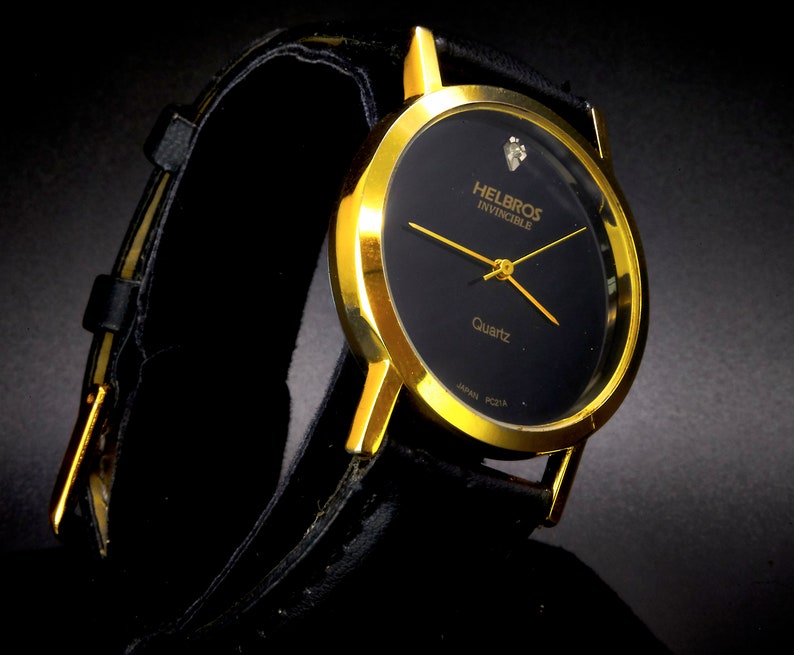 May include: A gold-tone wristwatch with a black face and a black leather band. The watch face has the words "Helbros Invincible" and "Quartz" printed on it.