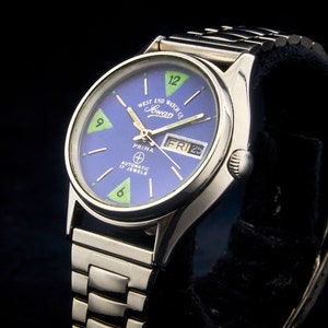 May include: A silver Prima automatic watch with a blue face and green accents. The watch face has the text "West End Watch Co. Sowar" and "Automatic 17 Jewels". The date window shows "FRI 25".