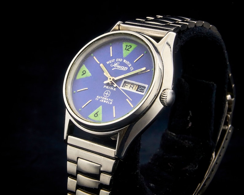 May include: A silver Prima automatic watch with a blue face and green accents. The watch face has the text "West End Watch Co. Sowar" and "Automatic 17 Jewels". The date window shows "FRI 25".