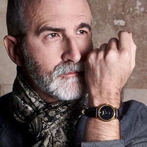 May include: A man wearing a gray blazer with a paisley patterned scarf. He is wearing a gold watch with a black face and a black leather band.