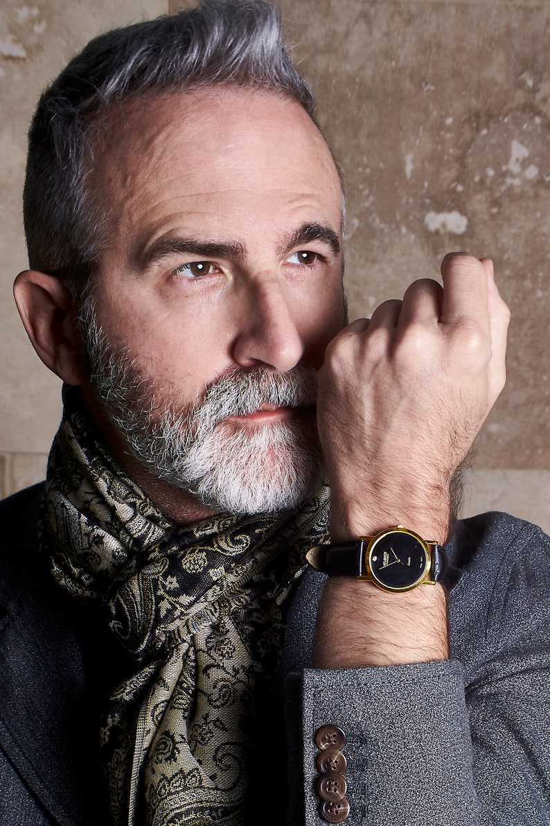 May include: A man wearing a gray blazer with a paisley patterned scarf. He is wearing a gold watch with a black face and a black leather band.