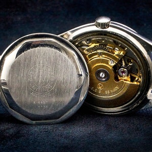 May include: A silver wristwatch with a stainless steel band. The watch has a round face and a silver case. The back of the watch is open, revealing the inner workings of the automatic movement. The watch is labeled "W.E.F. Automatic" and "Swiss Made".
