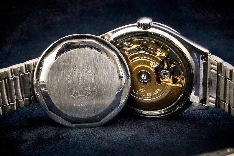 May include: A silver wristwatch with a stainless steel band. The watch has a round face and a silver case. The back of the watch is open, revealing the inner workings of the automatic movement. The watch is labeled "W.E.F. Automatic" and "Swiss Made".