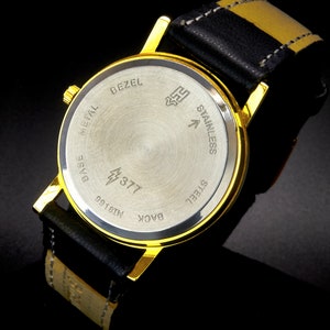 May include: The back of a gold-tone wristwatch with a black leather band. The watch face is made of stainless steel and has the words "Base Metal", "Bezel", "Back", "Stainless Steel", and "99181N" engraved on it. The watch also has a lightning bolt symbol and the number "377" engraved on it.