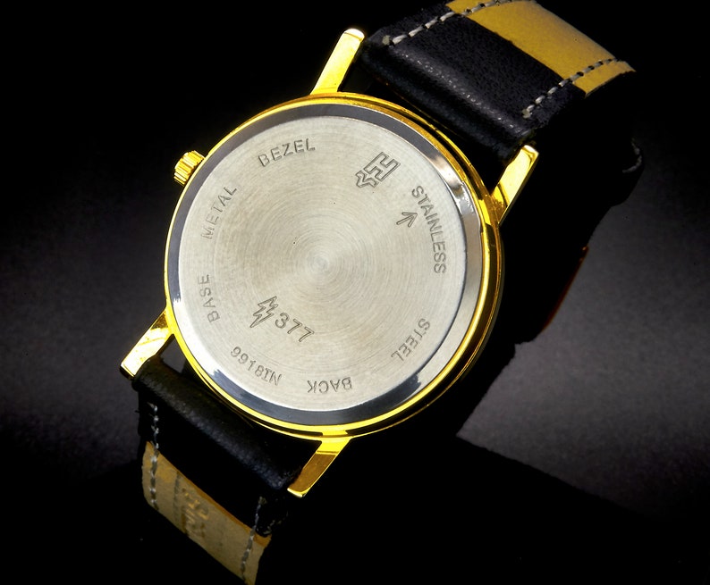 May include: The back of a gold-tone wristwatch with a black leather band. The watch face is made of stainless steel and has the words "Base Metal", "Bezel", "Back", "Stainless Steel", and "99181N" engraved on it. The watch also has a lightning bolt symbol and the number "377" engraved on it.