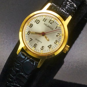 May include: A gold-toned Helbros Quartz watch with a silver face, black leather band, and gold hands. The watch face displays the numbers 1 through 12, with the brand name "Helbros" at the top. The watch band has a textured, crocodile-like pattern.