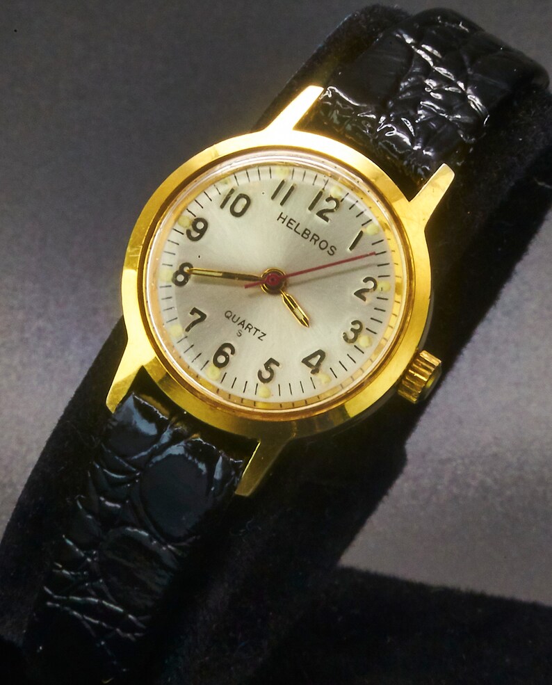 May include: A gold-toned Helbros Quartz watch with a silver face, black leather band, and gold hands. The watch face displays the numbers 1 through 12, with the brand name "Helbros" at the top. The watch band has a textured, crocodile-like pattern.