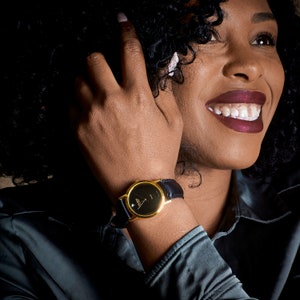May include: A gold-tone wristwatch with a black face and black leather band. The watch is on a person's wrist.
