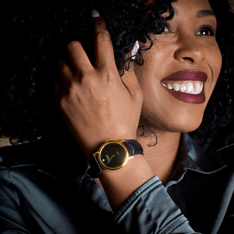 May include: A gold-tone wristwatch with a black face and black leather band. The watch is on a person's wrist.