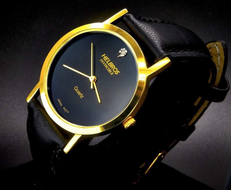 May include: A gold-tone wristwatch with a black face and a black leather band. The watch face has a small diamond at the 12 o'clock position and the words "Helbros Invincible Quartz" printed on it.