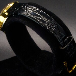 May include: A gold-toned wristwatch with a black leather band. The watch face is visible, and the band has a textured, crocodile-like pattern. The watch is displayed on a black surface, highlighting its design.