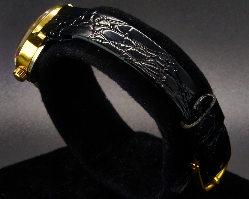 May include: A gold-toned wristwatch with a black leather band. The watch face is visible, and the band has a textured, crocodile-like pattern. The watch is displayed on a black surface, highlighting its design.