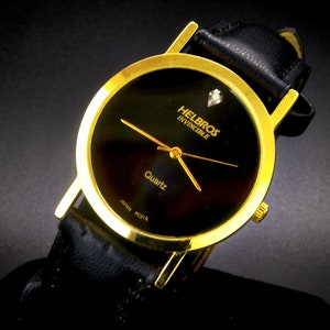 May include: A gold-tone Helbros Invincible quartz watch with a black face and a black leather band. The watch has a small diamond at the 12 o'clock position.