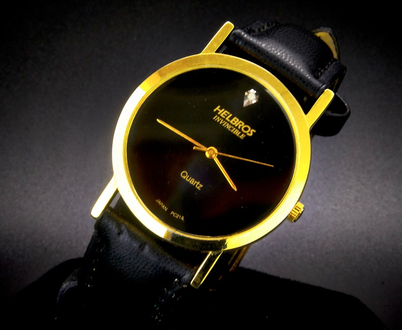 May include: A gold-tone Helbros Invincible quartz watch with a black face and a black leather band. The watch has a small diamond at the 12 o'clock position.