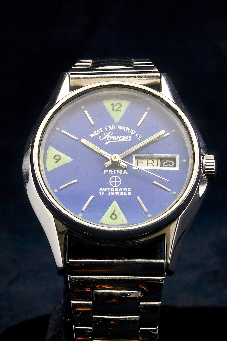 May include: A silver wristwatch with a blue face and white markings. The watch face has the text "West End Watch Co. Jowan Prima Automatic 17 Jewels" and the date "FRI 25".