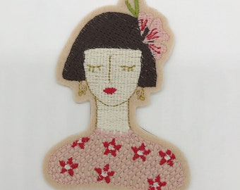 May include: Embroidered patch featuring a woman's portrait. The woman has dark brown hair, closed eyes, and red lips. She wears a pink dress with red flower accents and gold earrings. A pink flower adorns her hair.