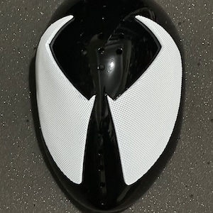 May include: A black and white mask with a glossy finish. The mask features a black base with white, wing-shaped accents around the eye area. The white sections have a mesh-like texture. Several small holes are visible on the mask's surface.