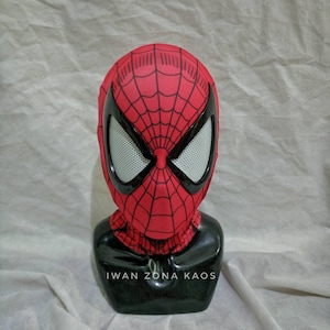 Op de afbeelding: Een rood en zwart Spider-Man-masker met een gaasoogbedekking. Het masker bevindt zich op een zwart mannequinhoofd met de tekst "Iwan Zona Kaos" op de basis.
