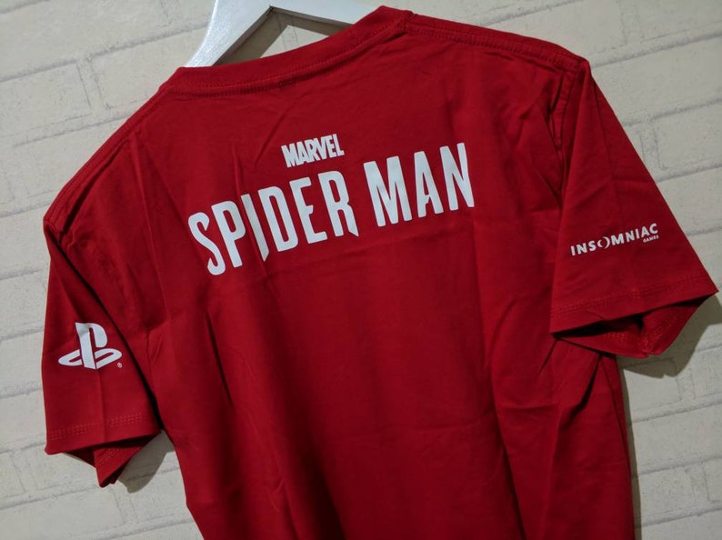 May include: Red t-shirt with white text that reads "MARVEL SPIDER-MAN" and "INSOMNIAC GAMES" on the back. The PlayStation logo is on the left sleeve.