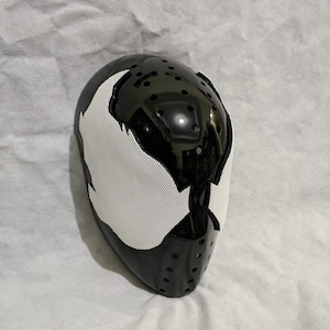 Op de afbeelding: Een zwart-wit masker met een glanzende afwerking. Het masker heeft een wit gaas oog- en mondgedeelte, met een zwart, geperforeerd onderste gedeelte. Het ontwerp doet denken aan een stripfiguur.