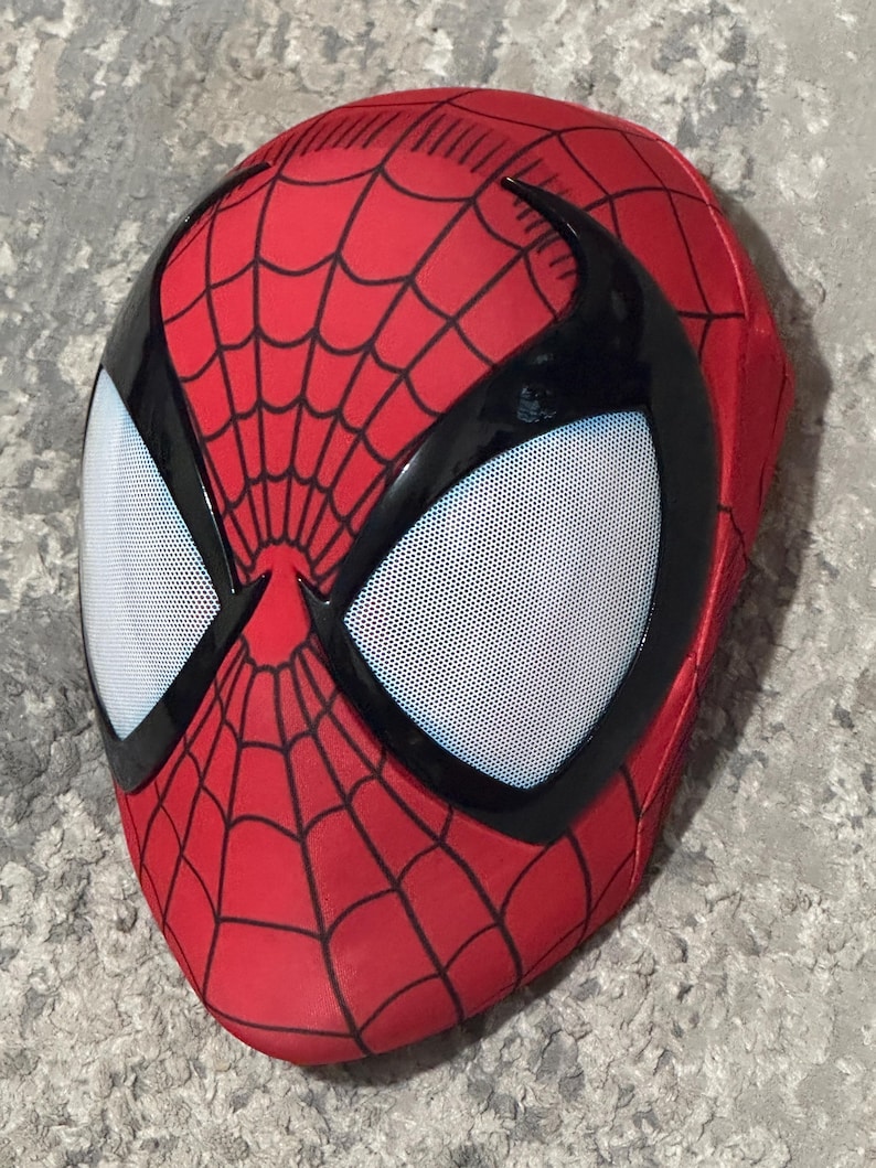 Op de afbeelding: Een rood Spider-Man-masker met een zwart spinnenwebpatroon en zwarte randen rond de ogen. De ooglenzen zijn wit met kleine, gelijkmatig verdeelde gaatjes. Het masker bedekt het hele gezicht.
