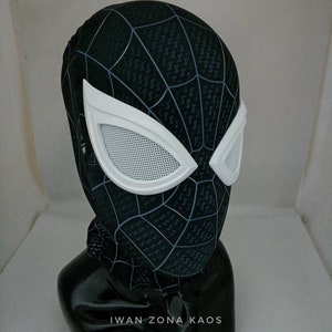 May include: Black and white Spider-Man mask with a white mesh eye piece. The mask has a black and white web pattern. The mask is on a black mannequin head. The text "Iwan Zona Kaos" is on the base of the mannequin.