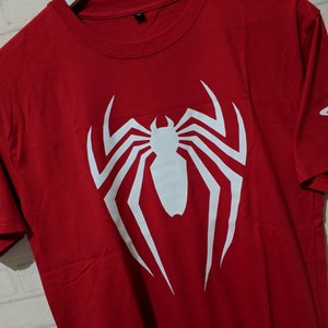 May include: Red t-shirt with a white spider-man logo and the text "INSOMNIAC GAMES" on the left sleeve and a PlayStation logo on the right sleeve.
