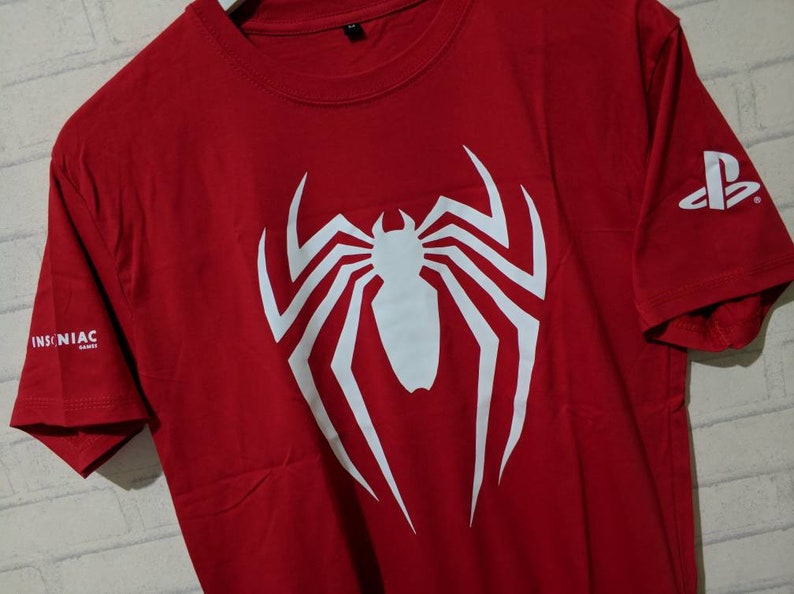 May include: Red t-shirt with a white spider-man logo and the text "INSOMNIAC GAMES" on the left sleeve and a PlayStation logo on the right sleeve.