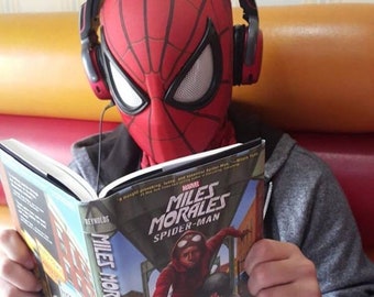 May include: A young person wearing a red and black Spider-Man mask and headphones is reading a Marvel Miles Morales Spider-Man graphic novel. The book cover features a red and black illustration of Miles Morales in a superhero pose.