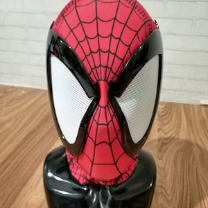 Op de afbeelding: Een zwarte en rode Bluetooth-luidspreker in de vorm van een Spider-Man-hoofd. De luidspreker heeft een zwarte basis en een rood-zwart Spider-Man-masker met een wit gaas dat de ogen bedekt.