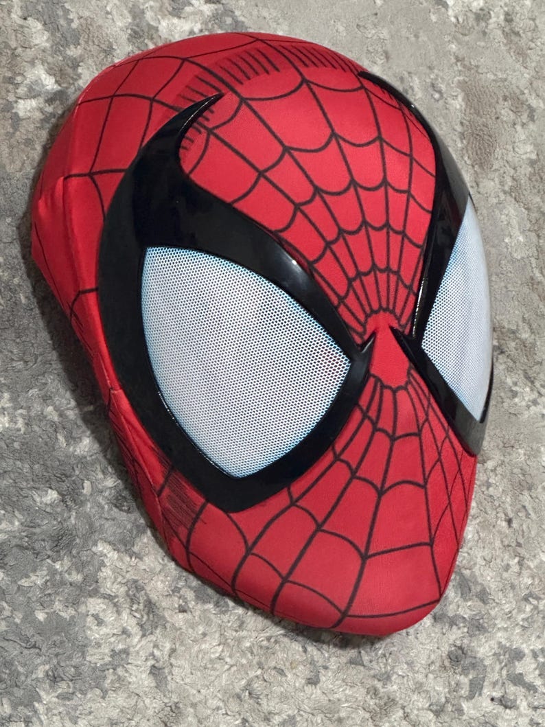 Op de afbeelding: Een rood Spider-Man masker met een zwart webpatroon en zwarte randen rond de ogen. De oogopeningen zijn van wit gaas. Het masker bedekt het hele hoofd.