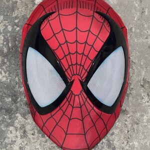 Op de afbeelding: Een rood Spider-Man masker met een zwart webpatroon en zwarte oogomrandingen. De oogopeningen zijn wit met kleine zwarte stippen. Het masker bedekt het hele gezicht.