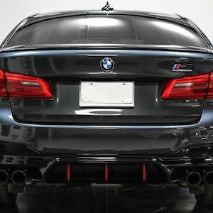 Może przedstawiać: Widok z tyłu czarnego BMW M5 z czarnym dyfuzorem tylnym i czterema końcówkami wydechowymi. Samochód ma czarne tablice rejestracyjne i logo BMW na bagażniku.