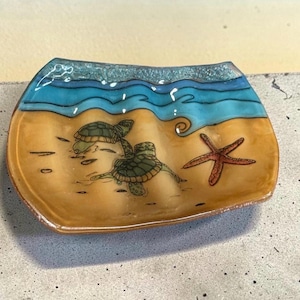 Könnte beinhalten: Eine rechteckige Glasschale mit einer Strandszene, zwei grünen Meeresschildkröten und einem roten Seestern auf beigem Sandgrund. Der obere Bereich zeigt blaue Meereswellen mit einem strukturierten, hellblauen Rand an der Oberkante der Schale.