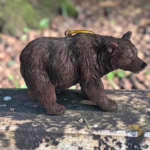Puede incluir: Una figura de oso pardo con detalles de pelaje texturizado, nariz negra y un pequeño lazo de color dorado unido a la parte superior de la espalda para colgar. El oso se muestra en posición de pie, mirando hacia el lado derecho del encuadre.