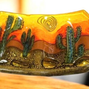 May include: A decorative glass dish with a desert landscape design. The dish features a vibrant orange and yellow sunset, with green cacti and a brown desert floor. A spiral sun is in the upper right corner. The dish has a curved shape.