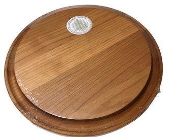 May include: A round wooden cutting board with a raised edge. The board is made of light brown wood and has a natural finish. The board is wrapped in plastic and has a sticker on it that says "Be Green,  Live Green,  Wood Products of America".